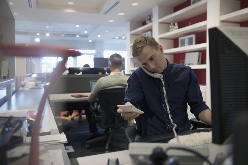 Businessman with calculator talking on telephone at computer in open plan office